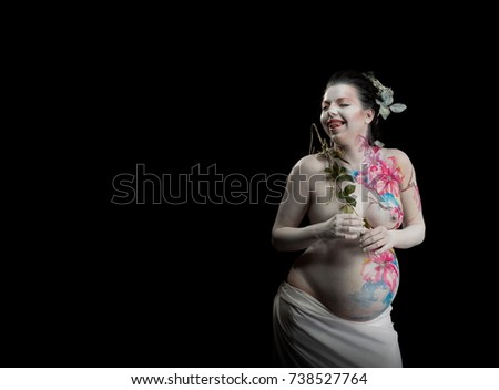 Emotional actress, pregnant woman with bodypainting on body and flowers orchid in hands posing on black background. White leather with large flowers and colored ornaments.