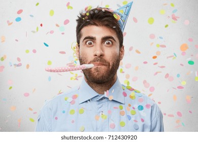 Emotional Adult Caucasian Male With Stubble Blowing Party Horn And Staring At Camera With Eyes Popped Out, Having Excited Look While Celebrating Birthday With Confetti Streamers Flying Around Him