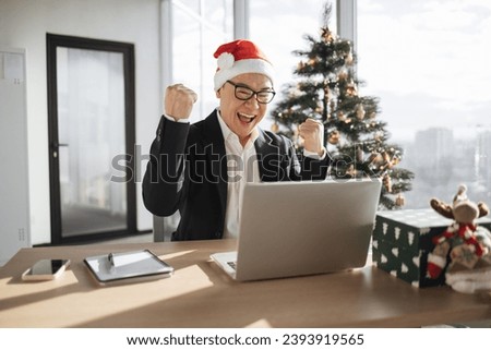 Emotional adult in Santa hat lifting fists of hands in excitement while looking at computer screen in office decorated with Christmas tree. Delighted asian male making successful holliday purchase