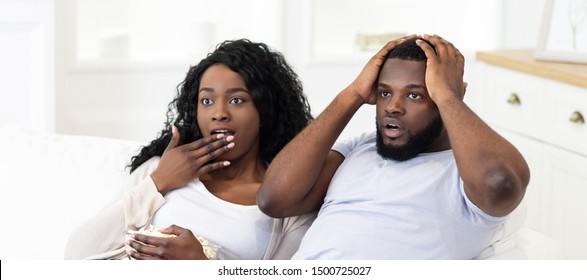 Emotional african american couple watching horror movie on television sitting on couch with popcorn, panorama