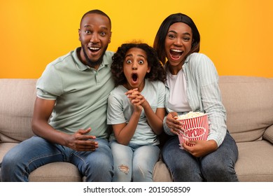 Emotional African American Family, Spouses With Daughter Eating Popcorn And Watching Movie On TV, Having Fun Together, Sitting On Couch Over Yellow Studio Background