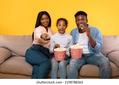 Emotional African American Family Of Three Watching Movie, Young Parents Sitting With Daughter On Couch, Eating Popcorn From Buckets, Woman Holding Remote Control, Yellow Studio Background