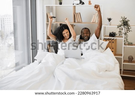 Emotional african american family of two raising hands in excitement while sitting in bed with laptop on blanket and credit card in hand. Thrilled married couple making successful deal online at home.