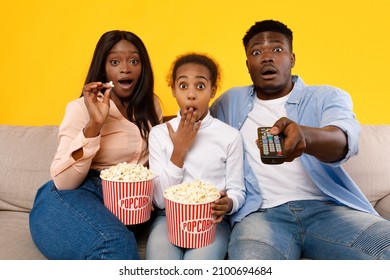 Emotional African American Family Watching Movie On TV And Eating Popcorn, Excited Spouses Sitting On Couch With Daughter And Covering Open Mouth Together, Isolated On Yellow Orange Studio Background