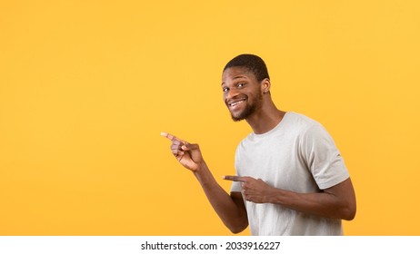 Emotional african american guy pointing fingers aside at empty space, posing and smiling over yellow studio background, panorama. Excited black man aiming at advertisement, indicating copy space