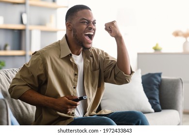 Emotional African American Man Watching Sports Game On TV Celebrating Victory Of Favorite Team Sitting On Sofa At Home. Sport Television Channel Programming Concept. Side View, Selective Focus