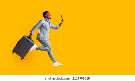 Emotional African American Millennial Guy In Casual Outfit With Suitcase Running Towards Copy Space, Holding Flight Ticket And Passport, Yellow Studio Background, Full Length Shot, Panorama, Side View