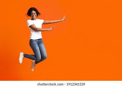 Emotionale afrikanische Jahrtausendfrau aus Amerika lächelt und weist auf Werbung oder Ankündigung hin. Aufgeregte schwarze Frau, die auf orangefarbenem Studiohintergrund springt und Kopienraum zeigt