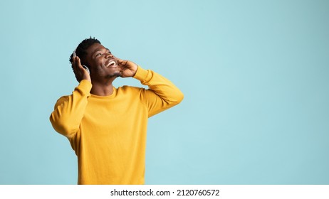 Emotional African American Millennial Man With Wireless Headset Looking At Copy Space For Advertisement And Smiling Over Blue Studio Background, Black Guy Listening To Nice Music