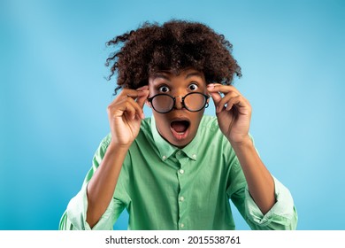 Emotional african american teen guy in glasses opening mouth in surprise, looking at camera over blue background. Black student looking overwhelmed and stunned, saying OMG or WOW