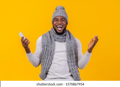 Emotional African American Winter Man Screaming Yes, Holding Cellphone And Raising Hands In The Air, Feeling Excited, Yellow Studio Background