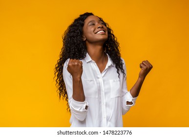 Emotional african american woman smiling and raising clenched fists in the air, feeling excited, yellow studio background