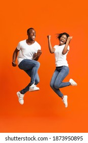 Emotional African American Young Couple Winners Clenching Fists And Screaming, Jumping Up Over Orange Studio Background. Happy Black Man And Woman Celebrating Success, Full Length Photo