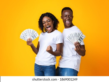 Emotional African-american Couple Holding Bunch Of Money Banknotes And Celebrating Success, Yellow Studio Background. Joyful Black Man And Woman Won Lottery, Showing Prize At Camera And Screaming