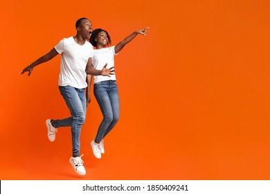 Emotional African-american Couple Jumping Up And Cheerfully Pointing Away At Empty Space, Orange Studio Background. Happy Young Black Man And Woman Aiming At Advertisement, Full Length Photo