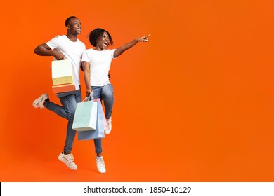 Emotional African-american Couple With Purchases Jumping Up And Cheerfully Pointing Away At Empty Space, Orange Studio Background. Happy Young Black Man And Woman Aiming At Advertisement