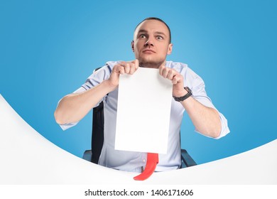 Emotional And Ambitious Young Leader Sits At The Table And Works With Documents And Papers. On A Blue Background Studio Photography