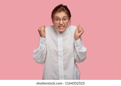 Emotional Angry Woman Clenches Fists With Vexed Expression, Cant Control Negative Emotions, Wears Round Spectacles, Isolated Over Pink Studio Wall. People, Bad Feeling And Body Language Concept