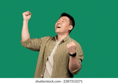 Emotional asian man shouting and shaking fists with eyes closed, celebrating big success over green studio background. Celebration of victory, joy emotion concept