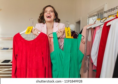 emotional attractive woman holding colorful dresses on hanger in clothing store, shopaholic, crazy exited emotion, happy face, spring summer fashion trend, choosing apparel, surprised discount sale