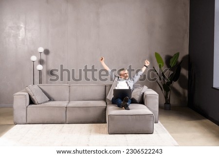 Emotional bearded middle-aged man celebrating success, gambling or trading online, using laptop while sitting on couch at home, copy space. Online gaming, financial markets concept