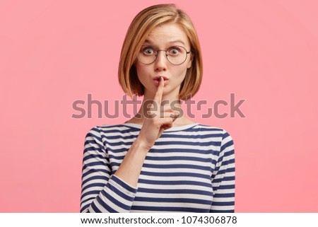 Emotional beautiful lovely woman with attractive appearance, keeps fore finger on lips, demonstrates silence gesture, ask to keep confidential information in secret, poses against pink wall.