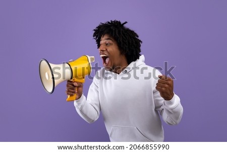 Emotional black adolescent shouting into megaphone, making announcement over violet studio background. Agitated African American teenager screaming with loudspeaker, advertising something