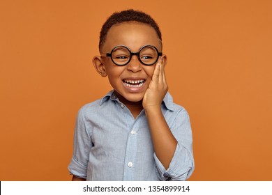 Emotional black dark-skinned little boy grimacing and holding hand on his cheek because of intolerable toothache. Handsome cheerful dark skinned African child in glasses and shirt smiling broadly