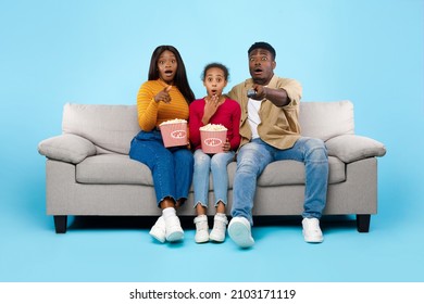 Emotional Black Family Watching Movie On TV And Eating Popcorn, Excited Spouses Sitting On Couch With Daughter Covering Open Mouth, Surprised Mom Pointing At Camera, Isolated On Blue Studio Background