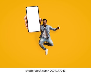 Emotional black guy in casual jumping up with brand new cellphone with blank screen, happy to use newest entertaining mobile application, mockup, orange studio background, copy space, full length