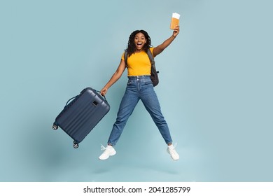 Emotional black lady in casual with backpack, luggage and passport, flight tickets going vacation, jumping in the air, sharing happiness, copy space, blue studio background. Vacation, trip, travelling