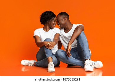 Emotional black lovers young man and woman cuddling on orange studio background, sitting on floor, touching each other with foreheads and laughing, enjoying time together, copy space