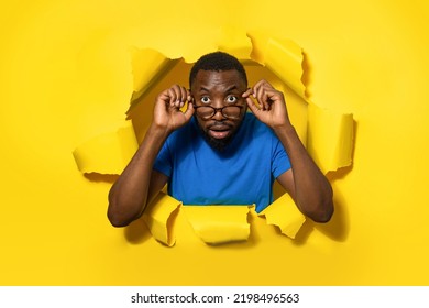 Emotional Black Man In Eyeglasses Looking At Camera Through Hole In Yellow Paper, Opening Mouth With Shock, Banner. Discount And Sale