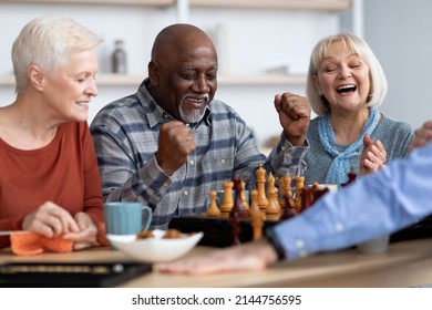 Emotional Black Man Playing Chess With Multiracial Friends, Happy Elderly Men And Women In Casual Outfits Sitting Around Table With Table Games, Chatting, Having Fun At Sanatorium