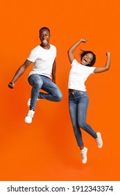 Emotional Black Millenial Couple Jumping Up On Orange Studio Background, Full Length Shot. Young Cheerful African American Man And Woman Celebrating Success, Having Fun Together