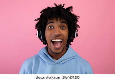 Emotional Black Teen Guy Listening To Popular Music In Headphones, Opening Mouth In Excitement On Pink Studio Background. Cool African American Adolescent Enjoying Cool Songs