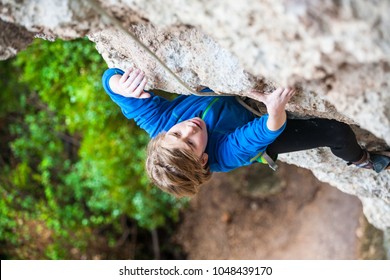 An Emotional Boy Climbs The Rock. The Child Is Engaged In Rock Climbing On A Natural Relief. The Sports Kid Actively Spends Time. Exercise In The Fresh Air. Rock Climbing On Difficulty.