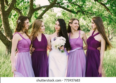 Emotional bride and bridesmaids are talking and smiling. Sexy caucasian girls in purple dresses having fun in the park, outdoors. Nature blurred background.