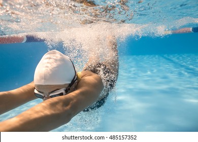 Emotional Close-up Photo Of Woman Who Swims Underwater In The Swimming Pool Outdoors. She Wears Dark Swimsuit, White Swim Cap And Swim Glasses. Sunlight Falls From Above. Splashes Are Around Her Body.