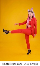 Emotional Cocky Model, Bold Person In Studio Making A Step Lifting Leg High. Young Woman In Red Suit Moving, Walking Movement. She Has Colorful Hair With Locks Afro Style, Braids Dreads Dreadlocks.