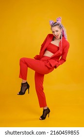 Emotional Cocky Model, Bold Person In Studio On Yellow Background. Daring Woman In Red Suit Moving, Lifting Her Leg Up. She Has Colorful Hair With Locks Afro Style, Braids Dreads Dreadlocks. 