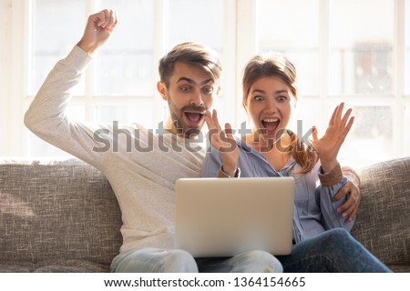 Emotional couple sitting on couch using laptop looking at device screen making big eyes open mouth feeling triumph received great news online lottery win, cheap flights tickets, huge discounts concept