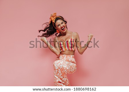 Emotional curly haired woman with red lipstick in striped bright top and pink cool trousers rejoices on isolated background..