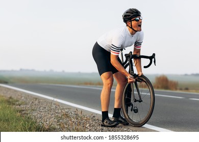 Emotional Cyclist In Helmet And Glasses Feeling Stress Of Broken Bike Somewhere At Countryside. Bearded Man Trying To Fix Wheel On Asphalt Road.