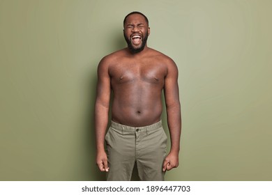 Emotional Dark Skinned Man With Naked Torso Wears Shorts Screams Loudly Opens Mouth Widely Has Thick Beard Poses Against Khaki Color Background Keeps Arms Down. Screaming Guy. Emotions Concept