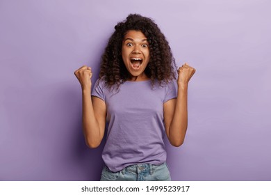 Emotional dark skinned woman with Afro hair, raises clenched fists, exclaims with excitemenet, rejoices sweet success, feels taste of victory, shouts for favourite team, wears purple t shirt and jeans