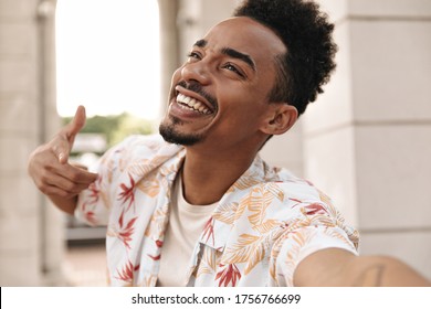 Emotional Excited Happy Man In Summer Floral Shirt Smiles Sincerely. Stylish Guy Points At Camera And Takes Selfie Outside.