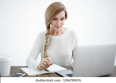 Emotional Excited Mature Woman With Long Braid Waiting At Desk With Empty Copybook, Smart Phone And Tea In Paper Cup, Reading Good News Online, Looking At Laptop Screen, Raising Her Eyebrows