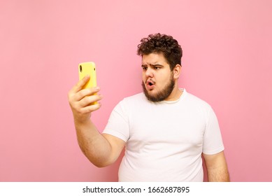 Emotional Fat Young Man In White T-shirt Is Using Smartphone On Pink Background, With Shocked Face Looking Into Camera. Funny Full Man Communicates On Video Call With Surprised Face.