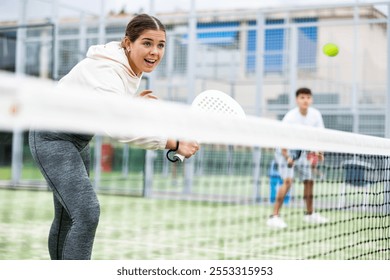 Emotional fit young female paddle tennis player waiting to receive serve, ready to perform backhand to return ball to opponent field on outdoor court on warm day - Powered by Shutterstock